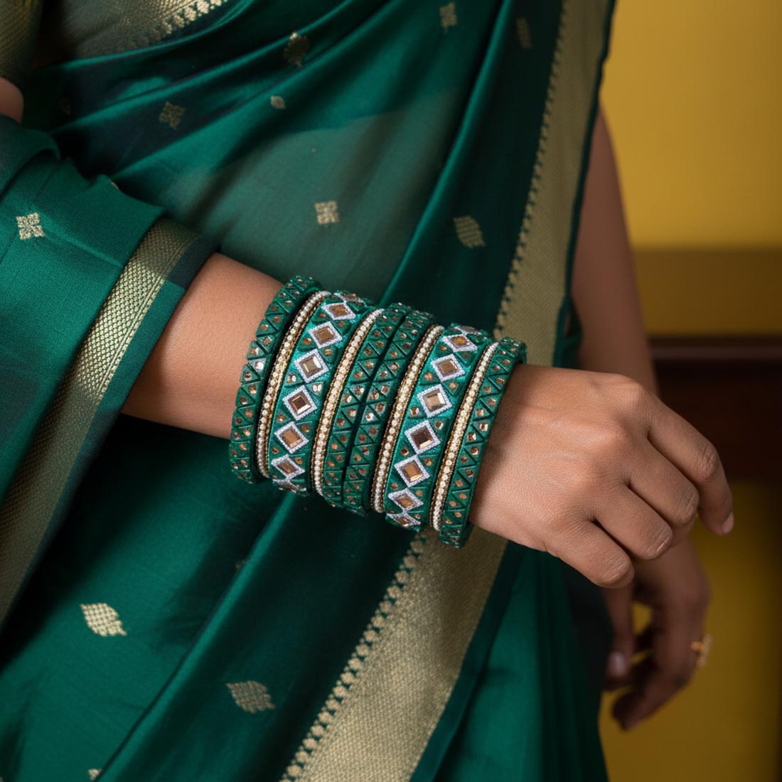 A traditional deep emerald green silk thread bangle set decorated with intricate geometric mirror work, tiny white seed beads, and gold diamond patterns, displayed stacked vertically on a rustic wooden slice surrounded by white baby's breath flowers and eucalyptus leaves.