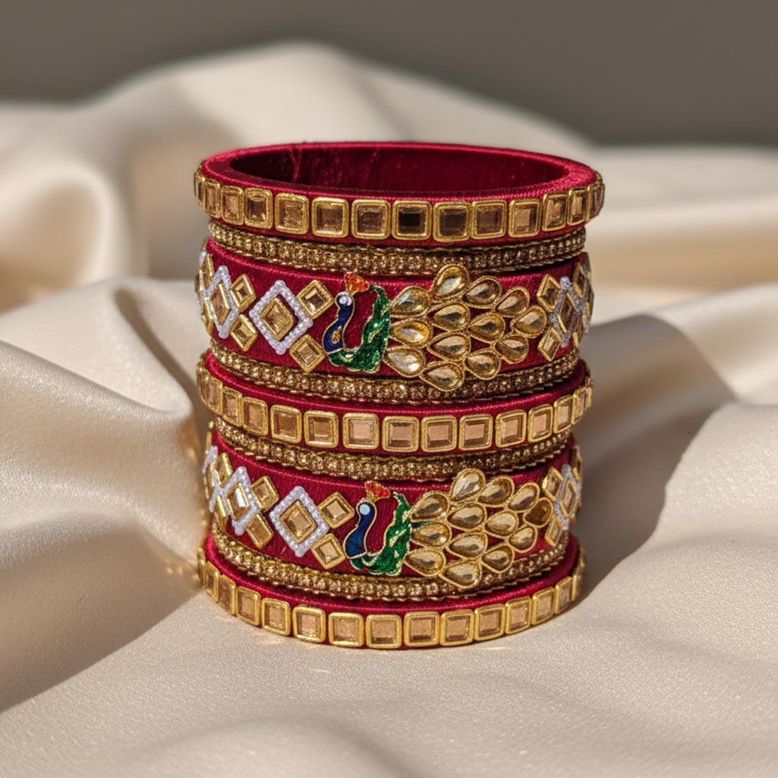 A royal maroon bridal bangle set wrapped in premium silk thread, beautifully decorated with gold-toned stone frames, geometric diamond rhinestone layouts, and dual enameled green and blue peacock motifs on a cream-colored silk backdrop.
