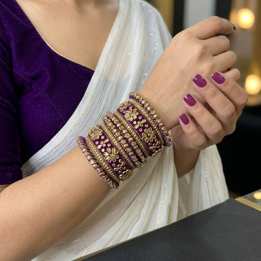 A royal purple silk thread bridal bangle set adorned with intricate gold floral Kundan work, shimmering rhinestone chain borders, and textured metallic accents, displayed stacked on a rustic wooden slice with draped purple fabric in the background.