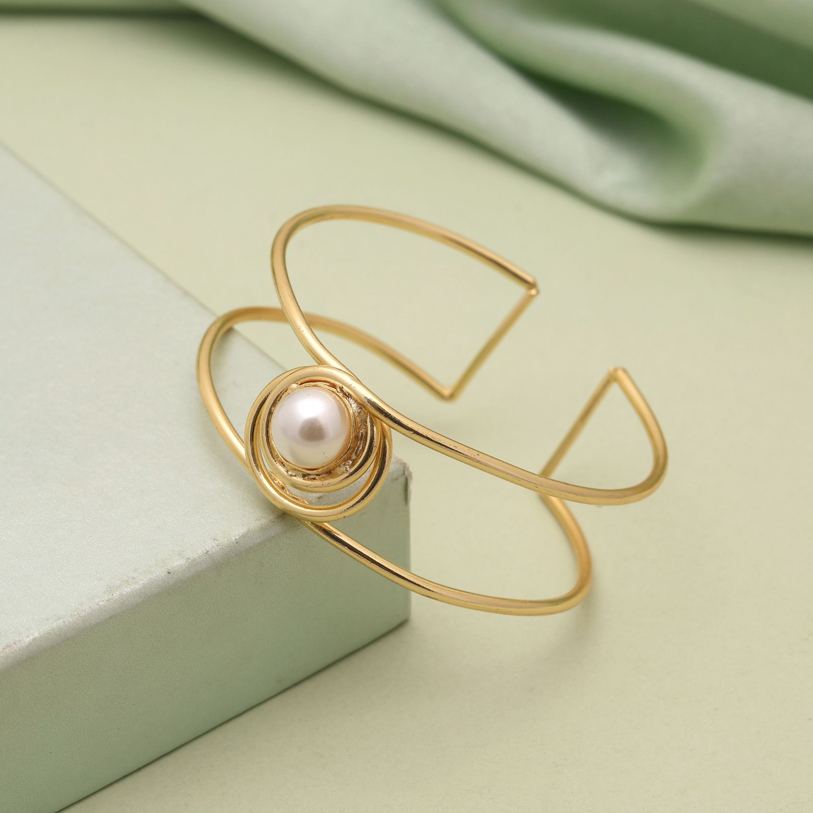 Gold-toned open cuff bracelet with two parallel wire bands and a central white pearl surrounded by concentric gold rings resting on textured dark wood.