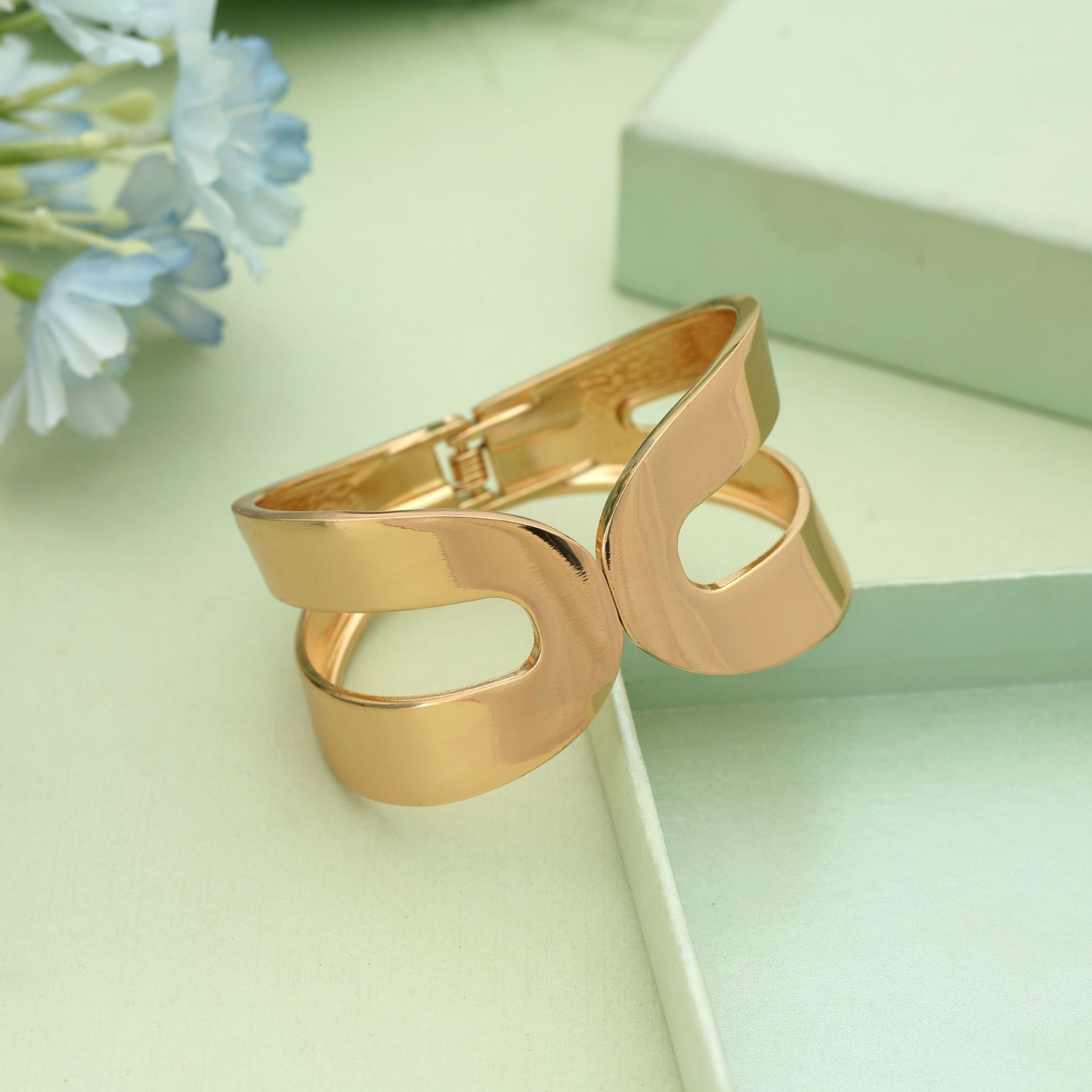 Gold-toned hinged cuff bracelet featuring a smooth mirror finish and an abstract dual-loop cutout design, displayed on a light gray surface with a gold box.