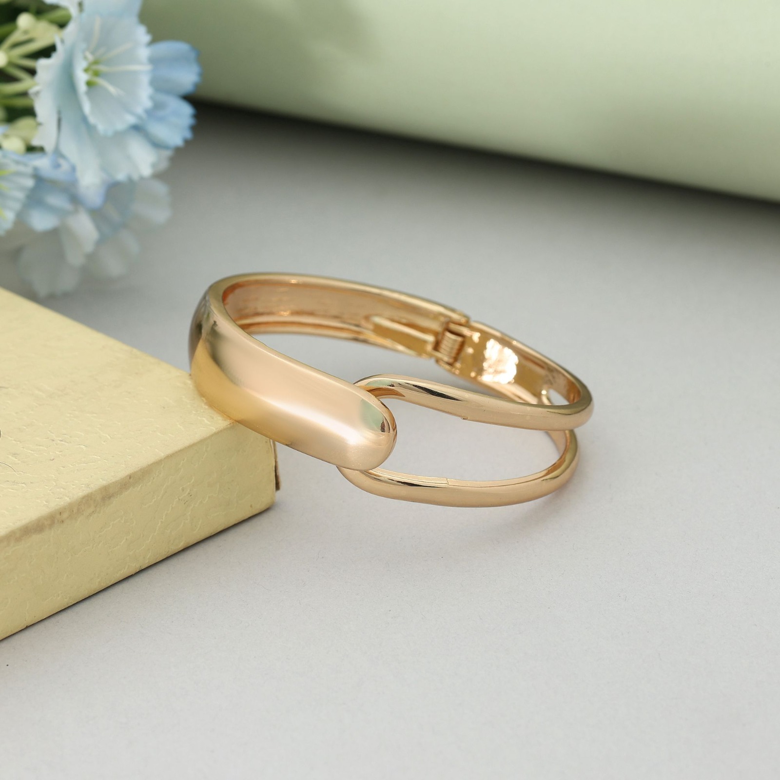 Gold-toned hinged cuff bracelet featuring an asymmetrical split-band design with a solid polished overlapping cap, resting on a soft green surface.