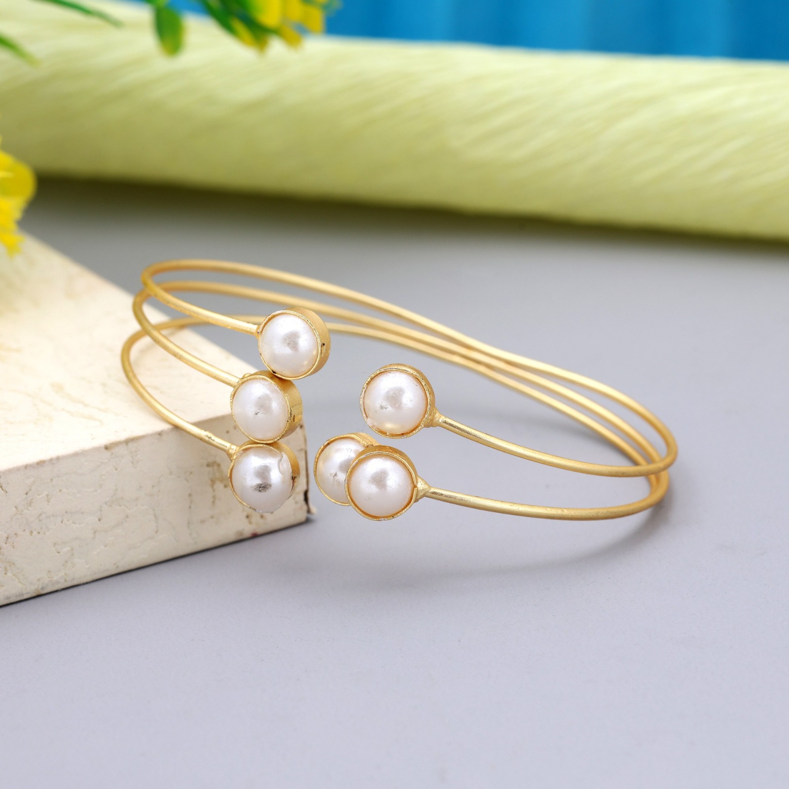Matte gold-toned triple wire open cuff bracelet featuring bezel-set white faux pearls on each open end, displayed on a gray surface.