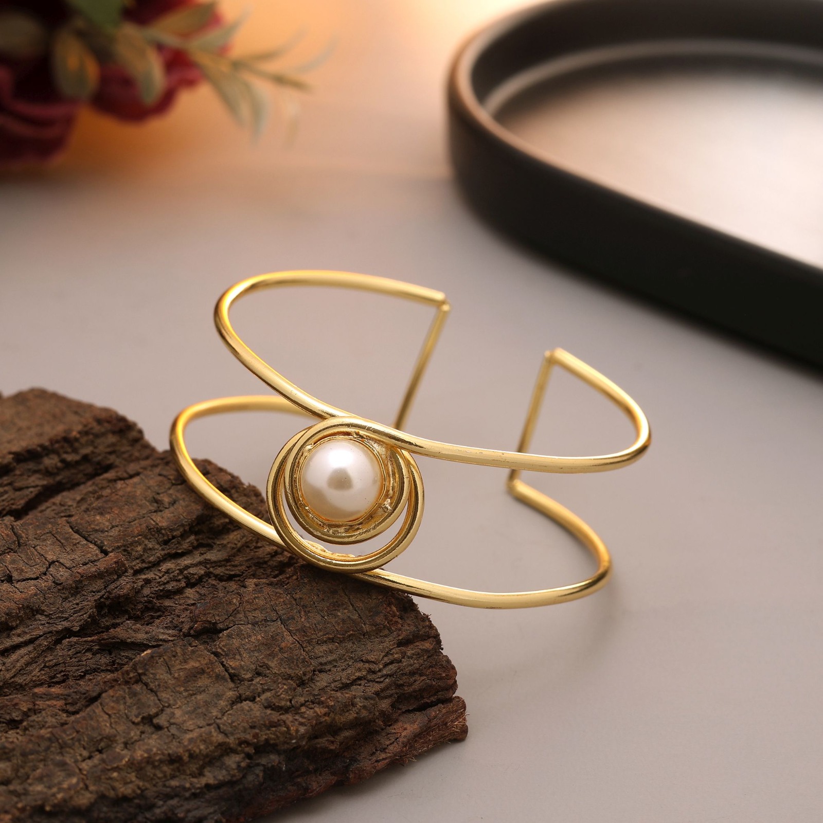 Gold-toned open cuff bracelet with two parallel wire bands and a central white pearl surrounded by concentric gold rings resting on textured dark wood.
