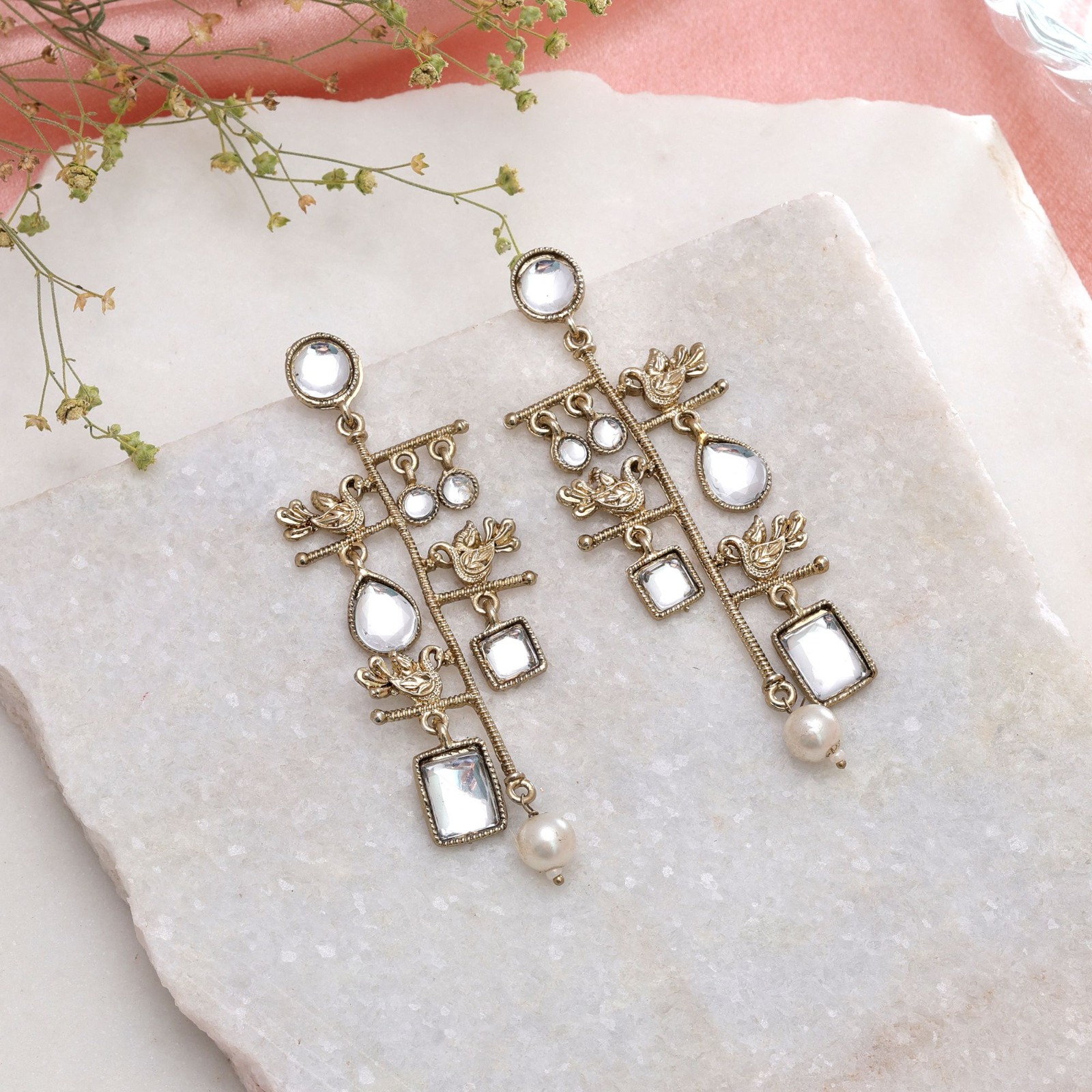 Pair of antique gold-toned linear drop earrings with geometric clear stones, small bird motifs, and a single pearl drop at the bottom, resting on a white marble surface.