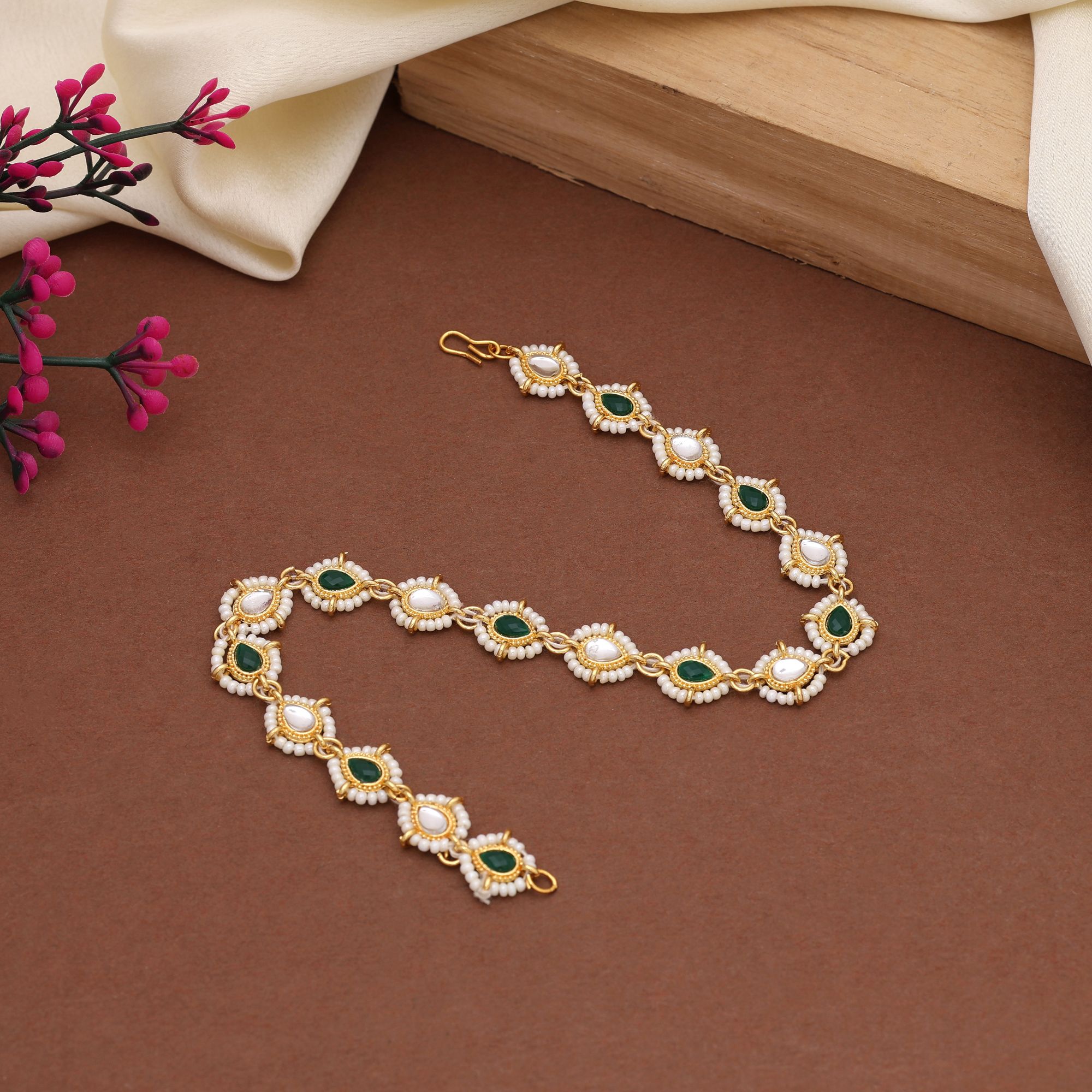 A traditional gold-plated Indian bridal matha patti hair chain featuring alternating teardrop-shaped emerald green stones and clear white Kundan crystals, framed by an intricate outline of tiny white seed beads and finished with a gold s-hook closure.