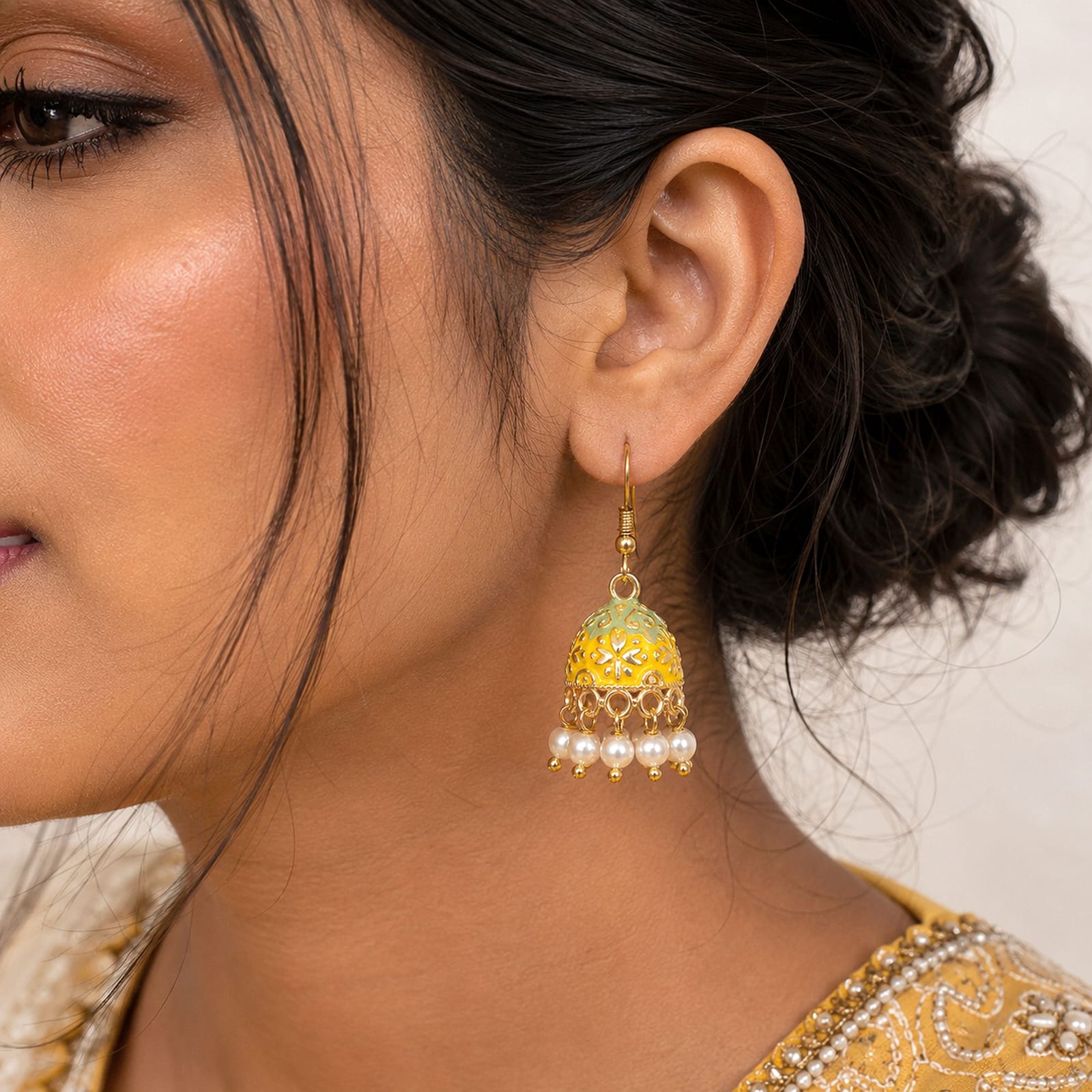 Add a touch of traditional charm to your ensemble with these gold-toned mini Meenakari jhumkas. Crafted with intricate enamel work, the bell-shaped dome highlights a vibrant dual-tone design of Mint Green at the top and Yellow at the base, detailed with intricate gold floral patterns. A delicate fringe of faux pearl drops along the bottom rim adds playful movement and elegance. Lightweight and fitted with classic French fish-hook closures, these earrings are perfect for festive occasions, ethnic celebrations, or elevating everyday traditional attire.