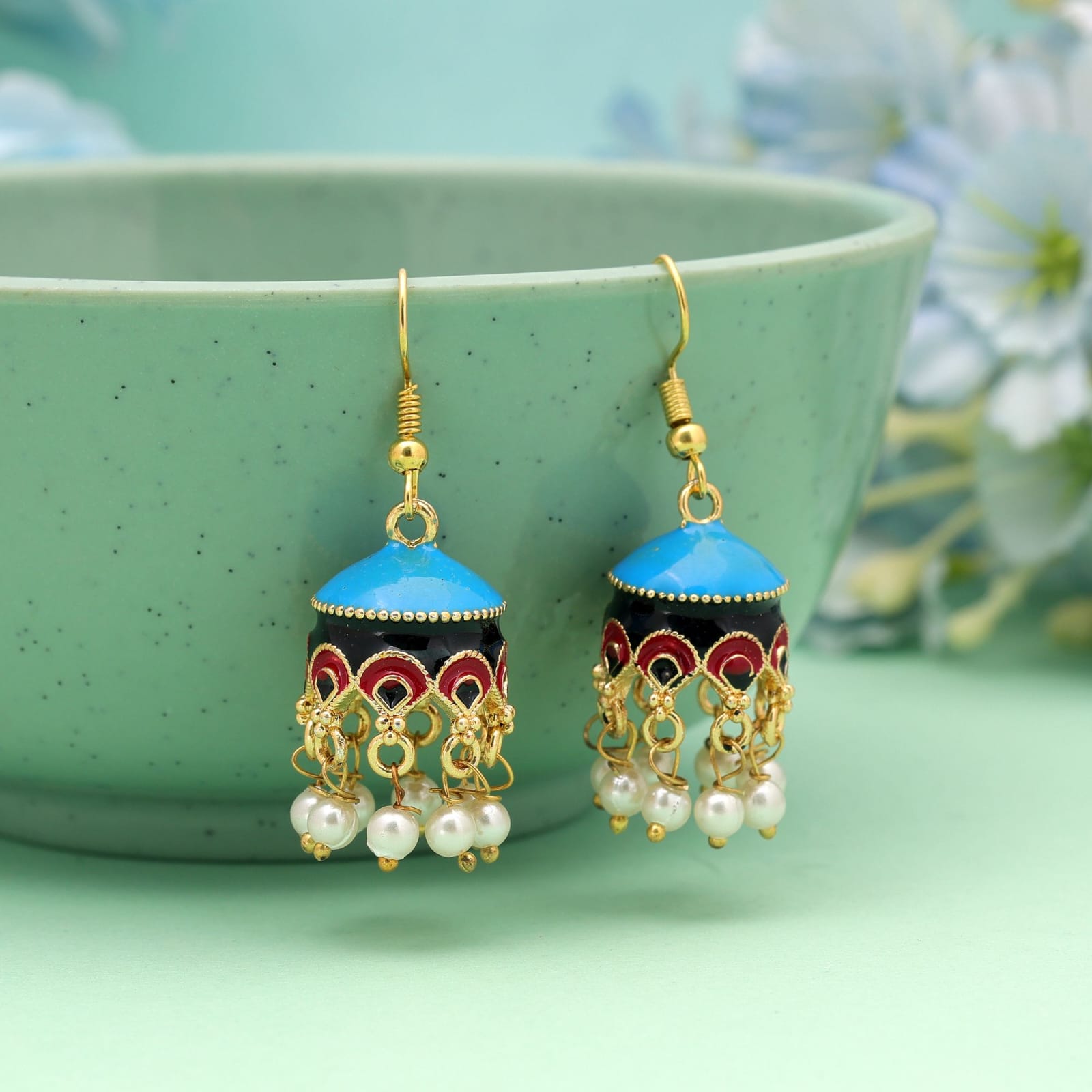 Elevate your ethnic wardrobe with these handcrafted Meenakari jhumka earrings. Designed with vibrant sky blue enamel tops and a contrasting black body adorned with intricate red and gold arch detailing, these lightweight bell-shaped earrings combine tradition with modern elegance. Finished with delicate dangling white pearl clusters, they offer an eye-catching movement that complements both festive attire and everyday traditional outfits. Key Features: Craftsmanship: Authentic Meenakari enamel handwork on a gold-toned base metal. Design: Dome-shaped jhumki with multicolored detailing and hanging faux-pearl beads. Closure: Easy-to-wear gold-plated fish hook ear wires. Occasion: Ideal for festive celebrations, weddings, cultural events, or casual ethnic wear. Styling Tip: Pair with pastel kurtis, festive sarees, or Indo-western fusion wear to let the sky blue hue pop.