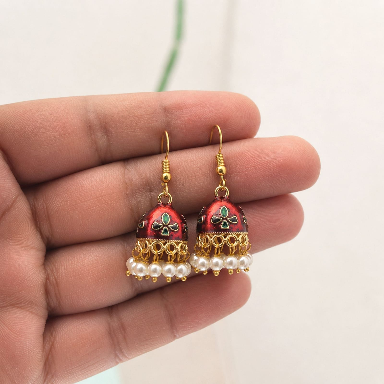 Bring subtle elegance to your wardrobe with these maroon Meenakari jhumka earrings, designed for both daily wear and special occasions. Crafted with a lightweight gold-toned frame, the compact bell-shaped domes feature smooth white enamelwork detailed with delicate red and green accents. A dainty rim of hanging white pearl beads adds soft charm and graceful movement. Styled with easy fish-hook earwires for all-day comfort, these versatile jhumkas pair effortlessly with everyday kurtas, casual fusion wear, or festive outfit ensembles.