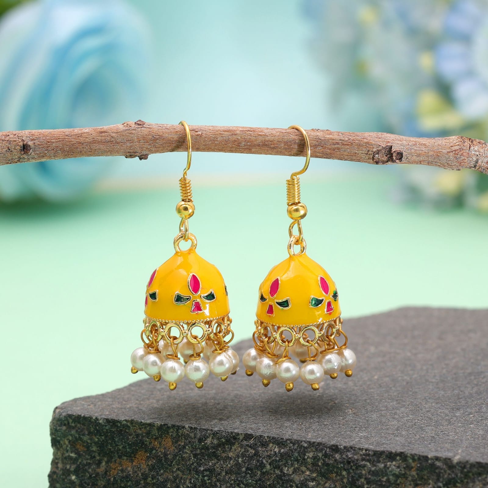 Bring subtle elegance to your wardrobe with these yellow Meenakari jhumka earrings, designed for both daily wear and special occasions. Crafted with a lightweight gold-toned frame, the compact bell-shaped domes feature smooth white enamelwork detailed with delicate red and green accents. A dainty rim of hanging white pearl beads adds soft charm and graceful movement. Styled with easy fish-hook earwires for all-day comfort, these versatile jhumkas pair effortlessly with everyday kurtas, casual fusion wear, or festive outfit ensembles.