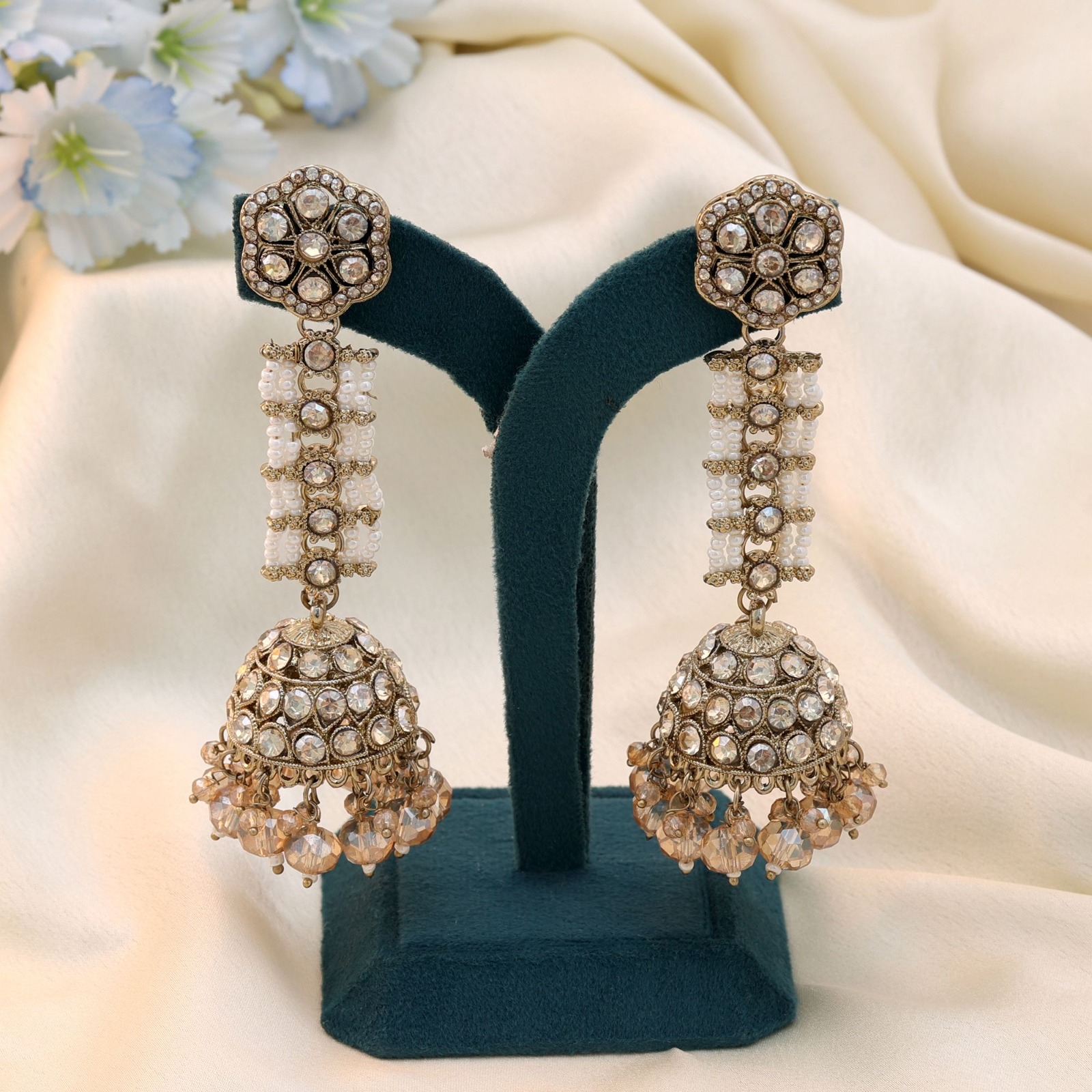 Antiqued gold statement jhumka earrings featuring a floral stud, white beaded multi-strand bridge, and crystal-studded dome with dangling champagne beads