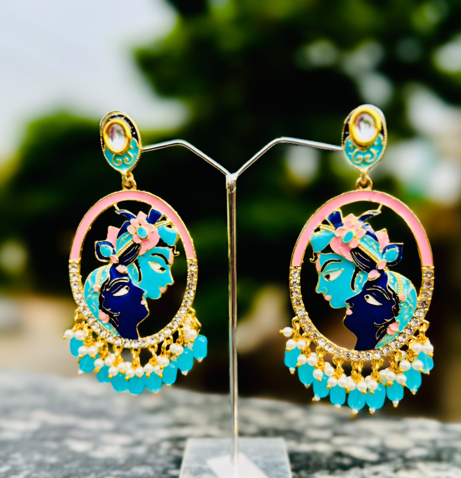 Pair of gold-tone Radha Krishna oval statement earrings with pink and turquoise enamel work, clear rhinestone accents, and a dangling fringe of small pearls and light blue teardrop beads.