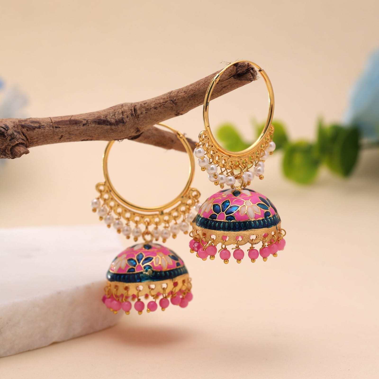 Add a vibrant pop of color to your ethnic wardrobe with these handcrafted Meenakari hoop jhumkas. Designed with a striking pink enamel dome featuring blue and white floral details, these statement earrings blend classic hoop charm with a traditional bell silhouette. Small white pearl accents trim the lower arch of the hoop, while matching pink beads dangle along the bottom edge for a graceful, eye-catching finish. Key Features: Craftsmanship: Detailed Meenakari handwork on a durable, gold-finished metal base. Design: Circular gold hoop structure paired with a floral-patterned jhumki dome and dangling pink beads. Closure: Standard hoop fastening for a lightweight, secure fit. Occasion: Perfect for festive celebrations, weddings, sangeet functions, and ethnic parties. Styling Tip: Style with a pink, royal blue, or white saree, Anarkali, or lehenga to highlight the dual-tone floral work.