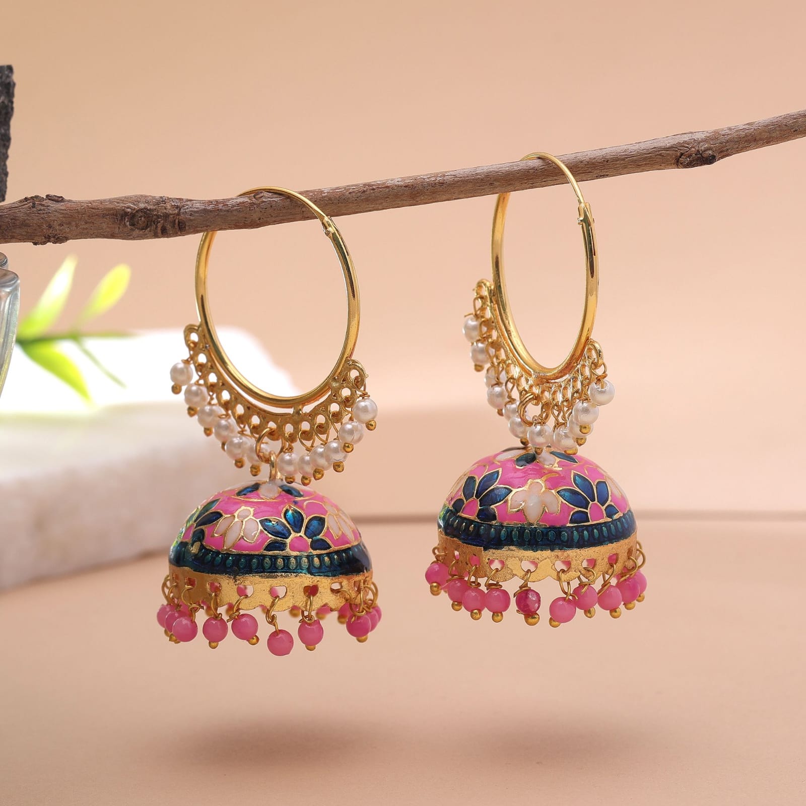 Add a vibrant pop of color to your ethnic wardrobe with these handcrafted Meenakari hoop jhumkas. Designed with a striking pink enamel dome featuring blue and white floral details, these statement earrings blend classic hoop charm with a traditional bell silhouette. Small white pearl accents trim the lower arch of the hoop, while matching pink beads dangle along the bottom edge for a graceful, eye-catching finish. Key Features: Craftsmanship: Detailed Meenakari handwork on a durable, gold-finished metal base. Design: Circular gold hoop structure paired with a floral-patterned jhumki dome and dangling pink beads. Closure: Standard hoop fastening for a lightweight, secure fit. Occasion: Perfect for festive celebrations, weddings, sangeet functions, and ethnic parties. Styling Tip: Style with a pink, royal blue, or white saree, Anarkali, or lehenga to highlight the dual-tone floral work.